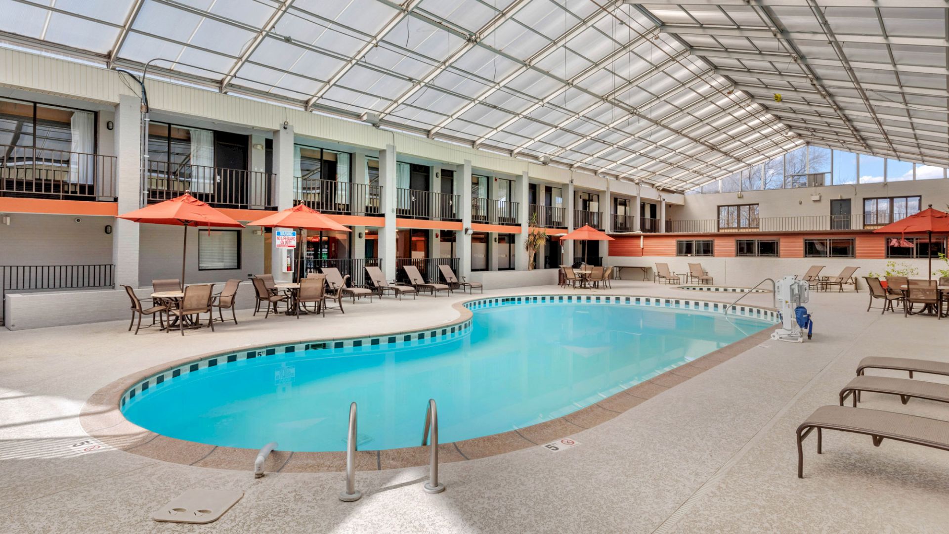 An indoor swimming pool with lounge chairs, tables, and orange umbrellas. The area is enclosed by a glass roof and surrounded by rooms.