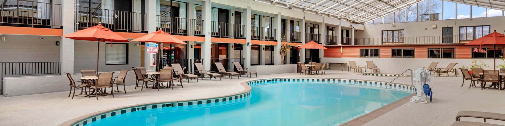 An indoor swimming pool with lounge chairs, tables, and orange umbrellas. The area is enclosed by a glass roof and surrounded by rooms.