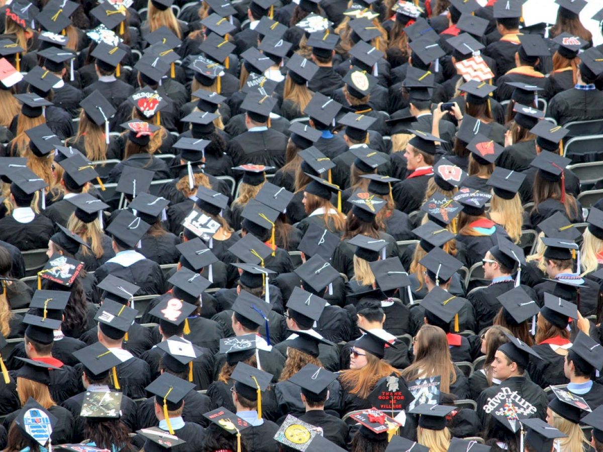 The image shows a large crowd of graduates wearing black caps and gowns, seated outdoors for a graduation ceremony.