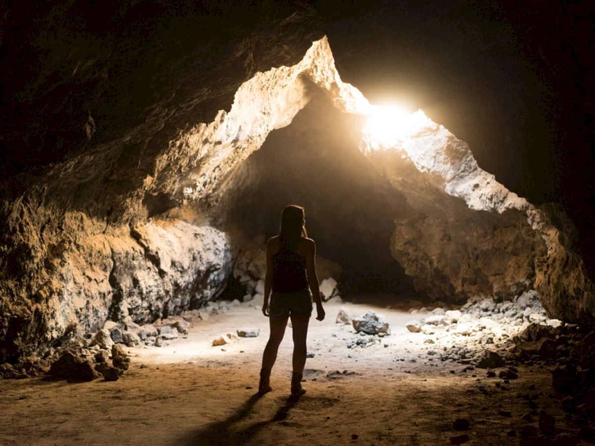 A person stands in a dimly lit cave, facing a bright opening ahead which illuminates parts of the rocky interior.