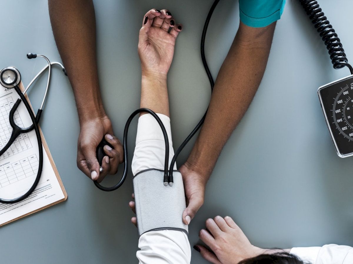 A healthcare professional measures a patient's blood pressure with a sphygmomanometer, as a stethoscope and clipboard rest nearby on the table.