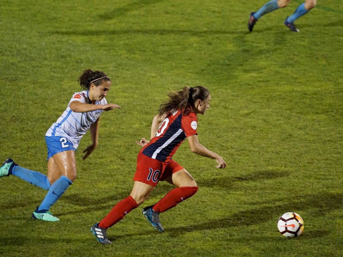 Two female soccer players are in action on the field; one in red and the other in blue, both running towards the soccer ball.