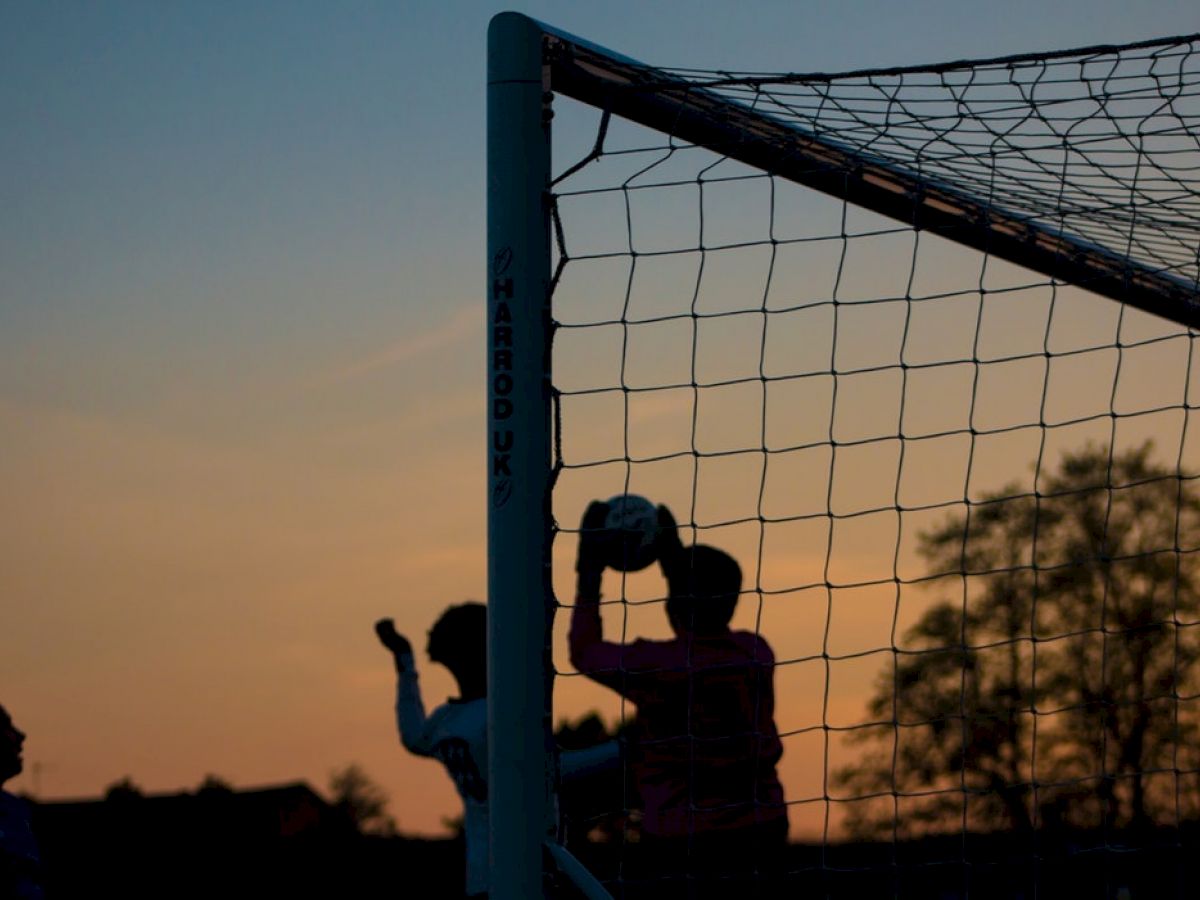 Silhouettes of people interacting near a soccer goal during sunset, with one person holding a ball.