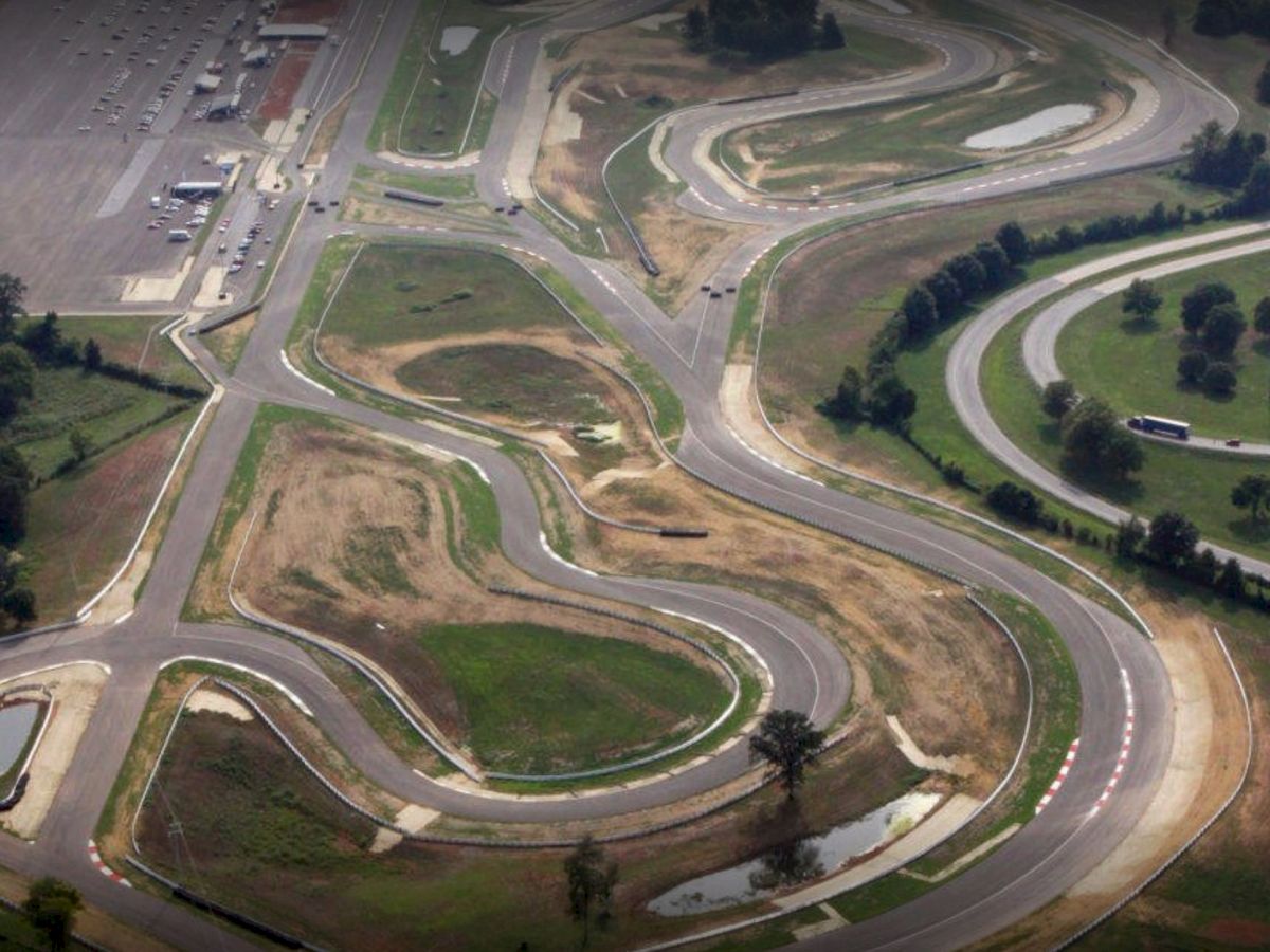 An aerial view of a winding racetrack with multiple curves, surrounded by greenery and parking areas.