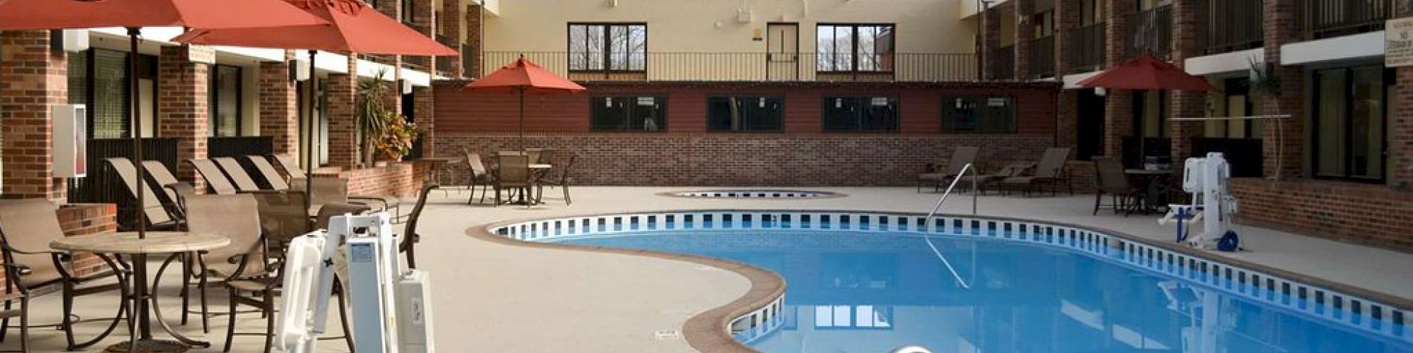 An indoor pool area with tables and red umbrellas, deck chairs, a pool lift, and a jacuzzi in the foreground.