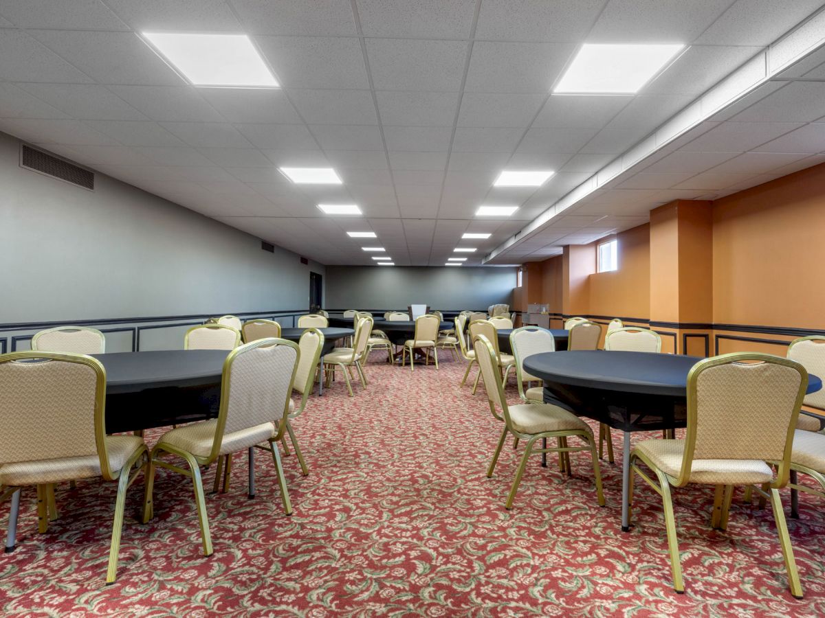 The image shows a conference or event room with several round tables and chairs arranged, a patterned carpet, and ceiling lights illuminating the space.