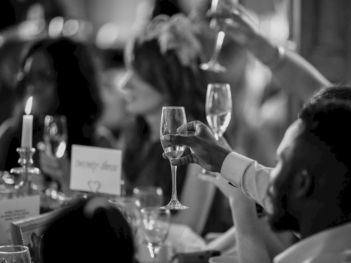 A group of people are raising their glasses in a toast at a formal event or celebration, with a candle and table settings in the foreground.