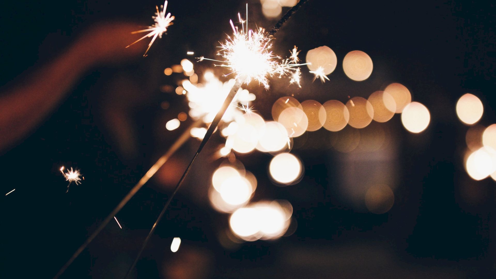 The image shows a close-up of sparklers being lit, with bright sparks and bokeh lights in the blurred background, creating a festive atmosphere.