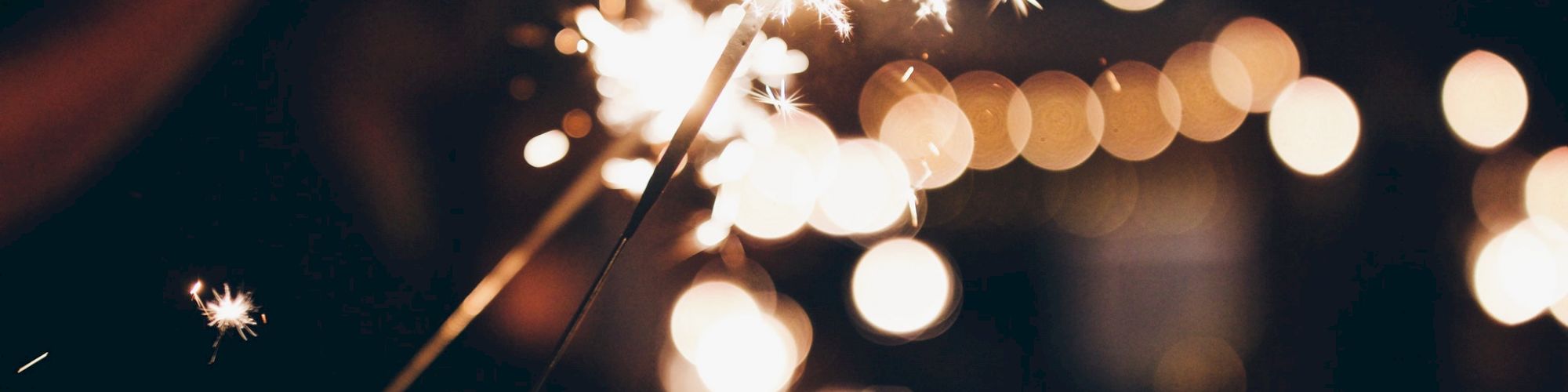 The image shows a close-up of sparklers being lit, with bright sparks and bokeh lights in the blurred background, creating a festive atmosphere.