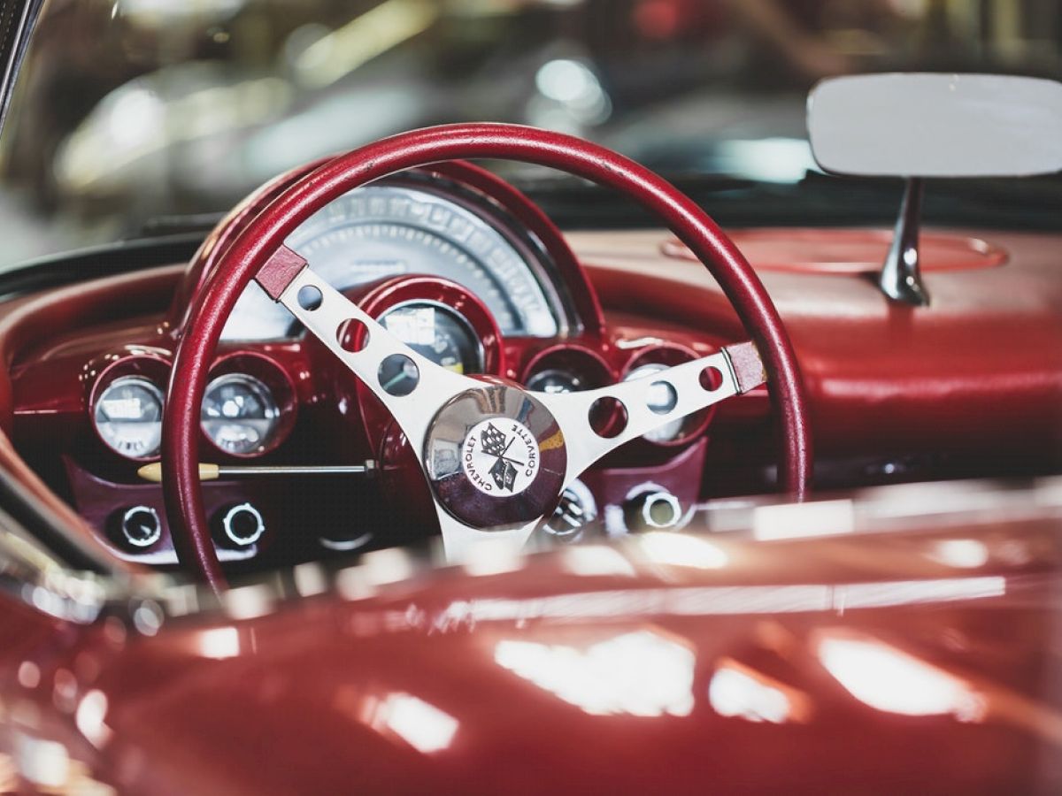 The image shows the interior of a vintage car with a red steering wheel, a classic speedometer, and glossy red details on the dashboard.