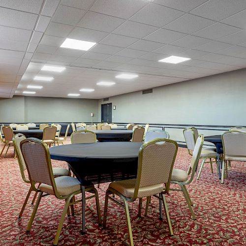 The image shows an empty, carpeted conference room with round tables and beige chairs under bright ceiling lights.