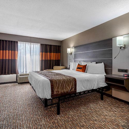 A contemporary hotel room features a king-sized bed, flat-screen TV, work desk, and window with curtains. Warm, neutral colors dominate the decor.