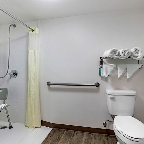 The image shows an accessible bathroom with a shower chair, grab bars, toilet, and a shelf holding towels and toiletries.