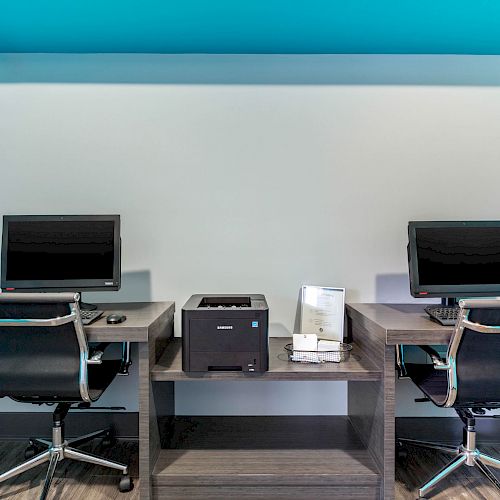 A small office setup with two computers, chairs, and a printer on a desk against a wall, surrounded by blue accents on the sides, ending the sentence.