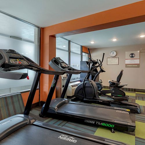 A gym room with treadmills, ellipticals, and a weight machine, featuring bright lighting and orange decor.