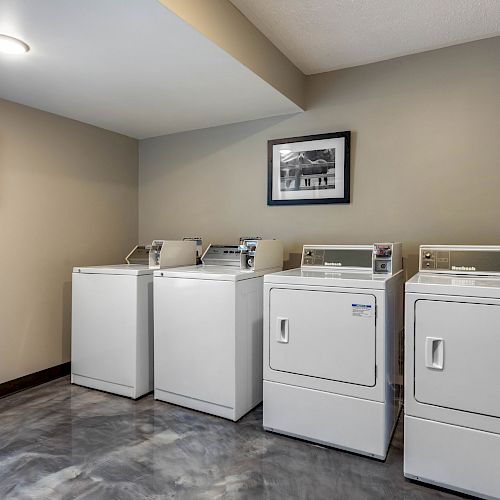 The image shows a laundry room with two washing machines and two dryers. There are framed pictures on the beige walls, and the floor is a polished gray.