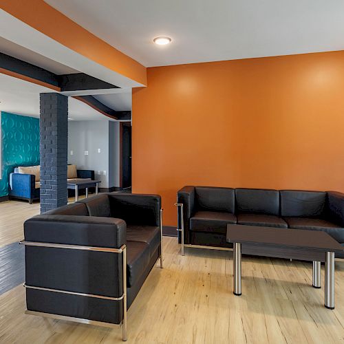 The image shows a modern living room with black sofas, orange and white walls, a wooden floor, and a small table with a lamp.