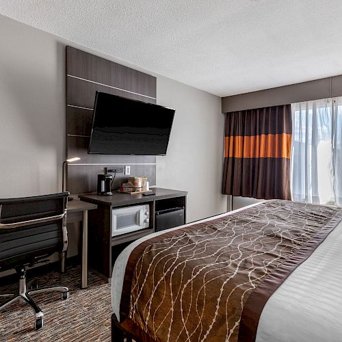 This image shows a hotel room with a neatly made bed, a desk with a chair, a wall-mounted TV, a microwave, a coffee maker, and large windows.