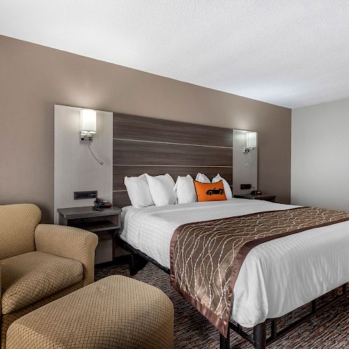 A modern hotel room with a king-sized bed, bedside tables, lamps, an armchair, and neutral decor. The bed has four pillows and a decorative pillow.