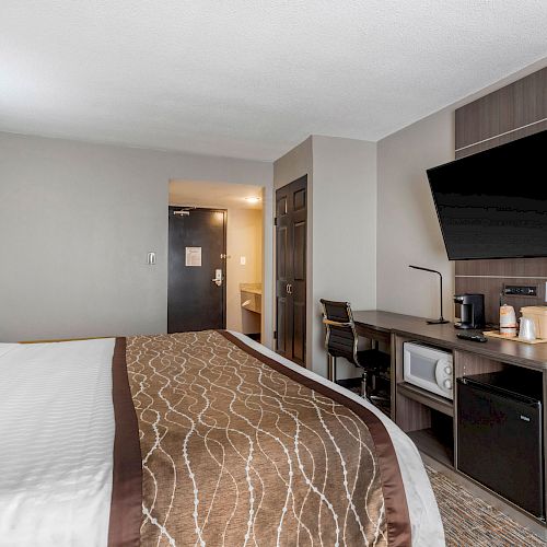 A modern hotel room featuring a large bed, a wall-mounted TV, a desk, a coffee maker, a microwave, and a mini-fridge, with a minimalist design.