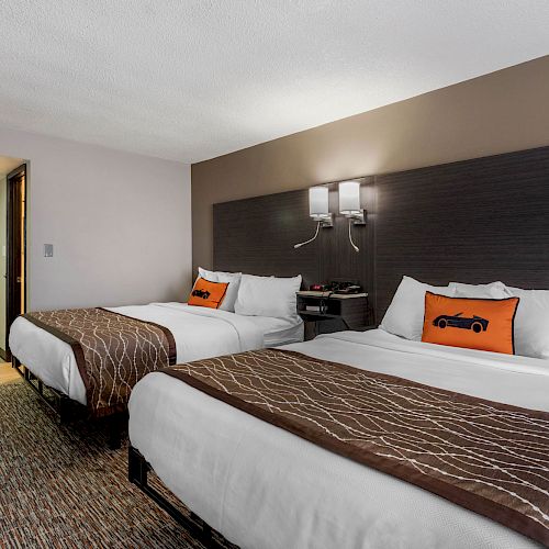 A hotel room with two large beds, brown and white bedding, a wall-mounted lamp, and a nightstand between the beds is visible.