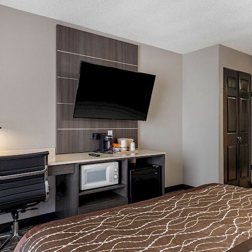 This image shows a hotel room with a wall-mounted TV, desk with lamp and chair, a microwave, mini-fridge, and a view of the bathroom area.