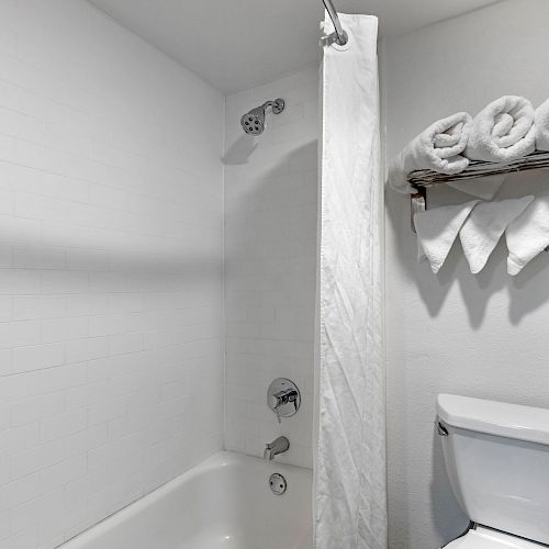 The image shows a clean bathroom with a white bathtub, a showerhead, a white curtain, a toilet, and shelves with neatly rolled and folded towels.