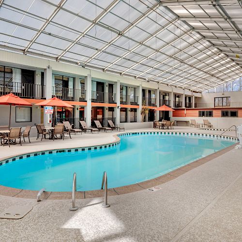 An indoor swimming pool area with lounge chairs, tables, and orange umbrellas. The ceiling is transparent, allowing natural light to come in.