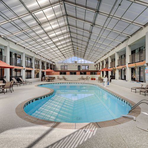 The image shows an indoor pool area with lounge chairs, tables with red umbrellas, and a glass ceiling allowing natural light to flood the space.