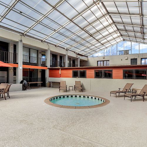 An indoor courtyard features a small pool, lounge chairs, tables with umbrellas, and a high ceiling with large windows allowing natural light to pour in.