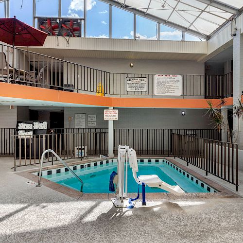 An indoor pool area with a blue chair lift, surrounded by railings, lounge chairs on the upper level, and signs on the walls.