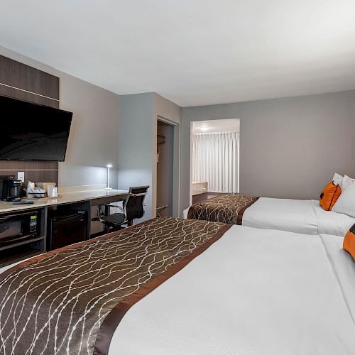 This image shows a modern hotel room with two double beds, a wall-mounted TV, a desk, a chair, and a coffee maker on the counter.