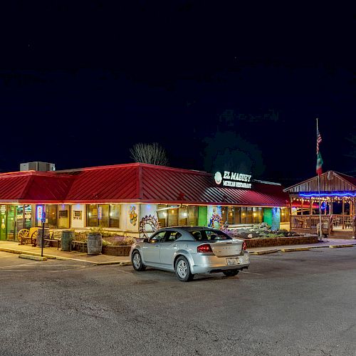 The image shows a brightly lit restaurant with a red roof in a parking lot at night, cars parked in front, and a festive gazebo decorated with lights.