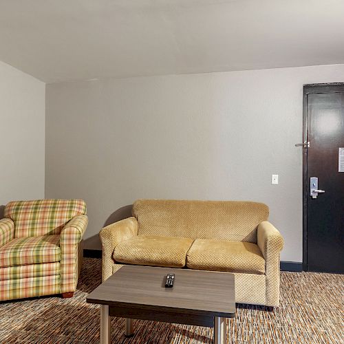 A small living area with a plaid armchair, beige sofa, coffee table, and a door slightly open revealing a hallway.