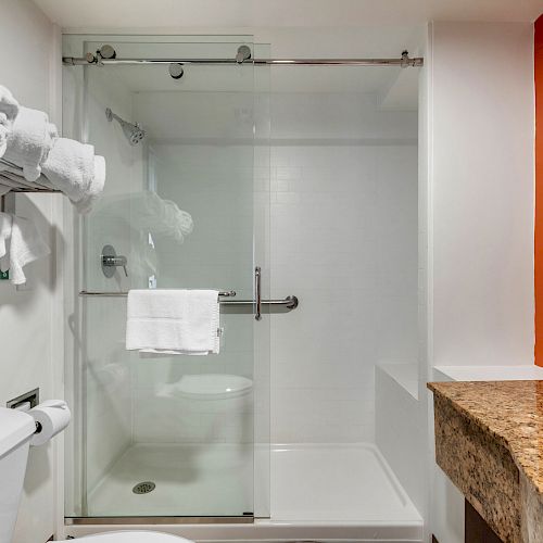 A modern bathroom with a glass-enclosed shower, granite countertop, and folded towels on a rack above a toilet.