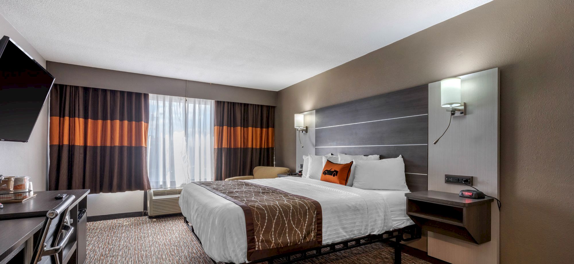 A modern hotel room with a king-sized bed, wall-mounted TV, desk, and two nightstands. The window has dark curtains with orange accents.