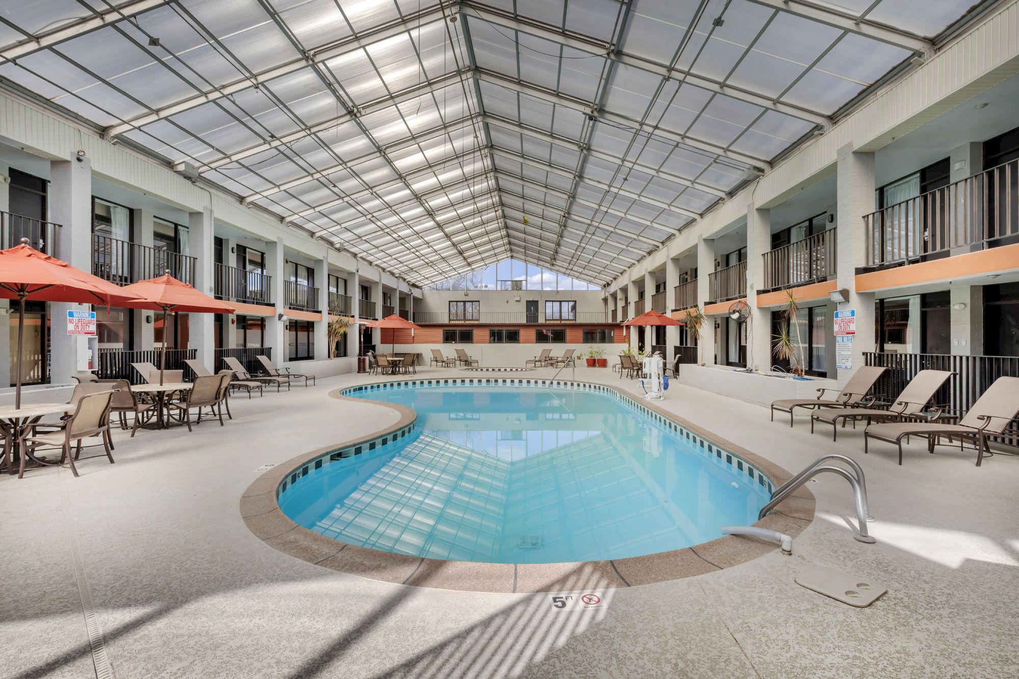 An indoor swimming pool area with lounge chairs, umbrellas, and a glass ceiling allowing natural light. The space appears clean and inviting.