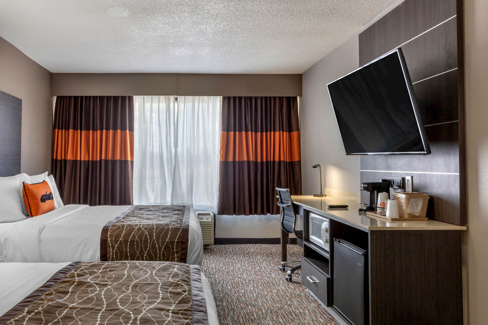 The image shows a hotel room with two beds, a window with curtains, a wall-mounted TV, a desk with a chair, and a mini-fridge.