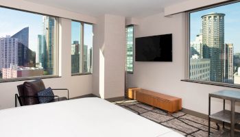 A modern hotel room with a city view, featuring large windows, a TV, a lounge chair, a wooden cabinet, and a side table.