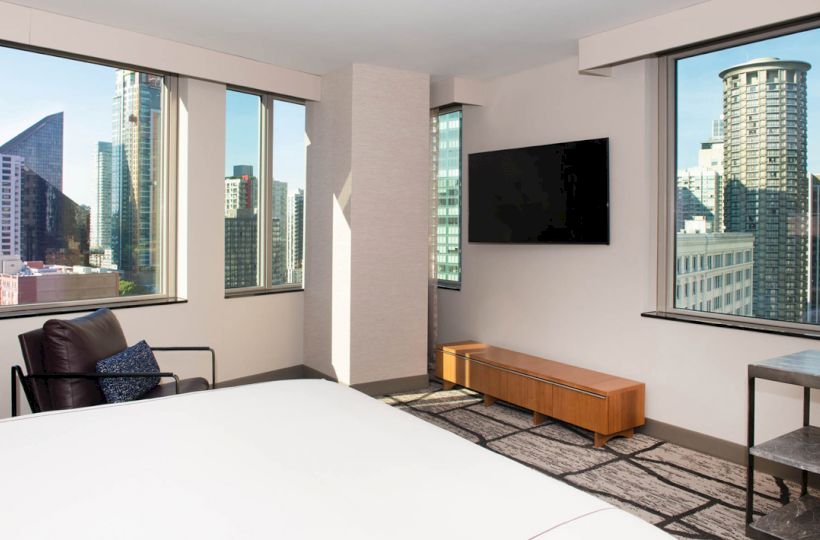 A modern hotel room with a city view, featuring large windows, a TV, a lounge chair, a wooden cabinet, and a side table.