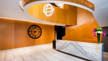 A modern lobby featuring a marble reception desk, a large wall clock, wood paneling, and stylish circular light fixtures with vibrant floral arrangements.