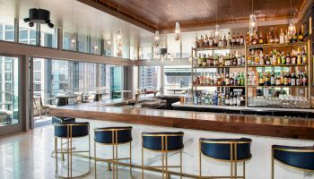 The image shows a modern bar with high chairs, a wooden countertop, and shelves stocked with various bottles, located in a high-rise building.