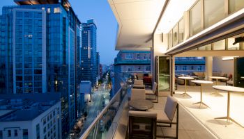 This image shows a modern balcony at dusk with tables and chairs, overlooking a city street bordered by tall buildings and soft evening lights.
