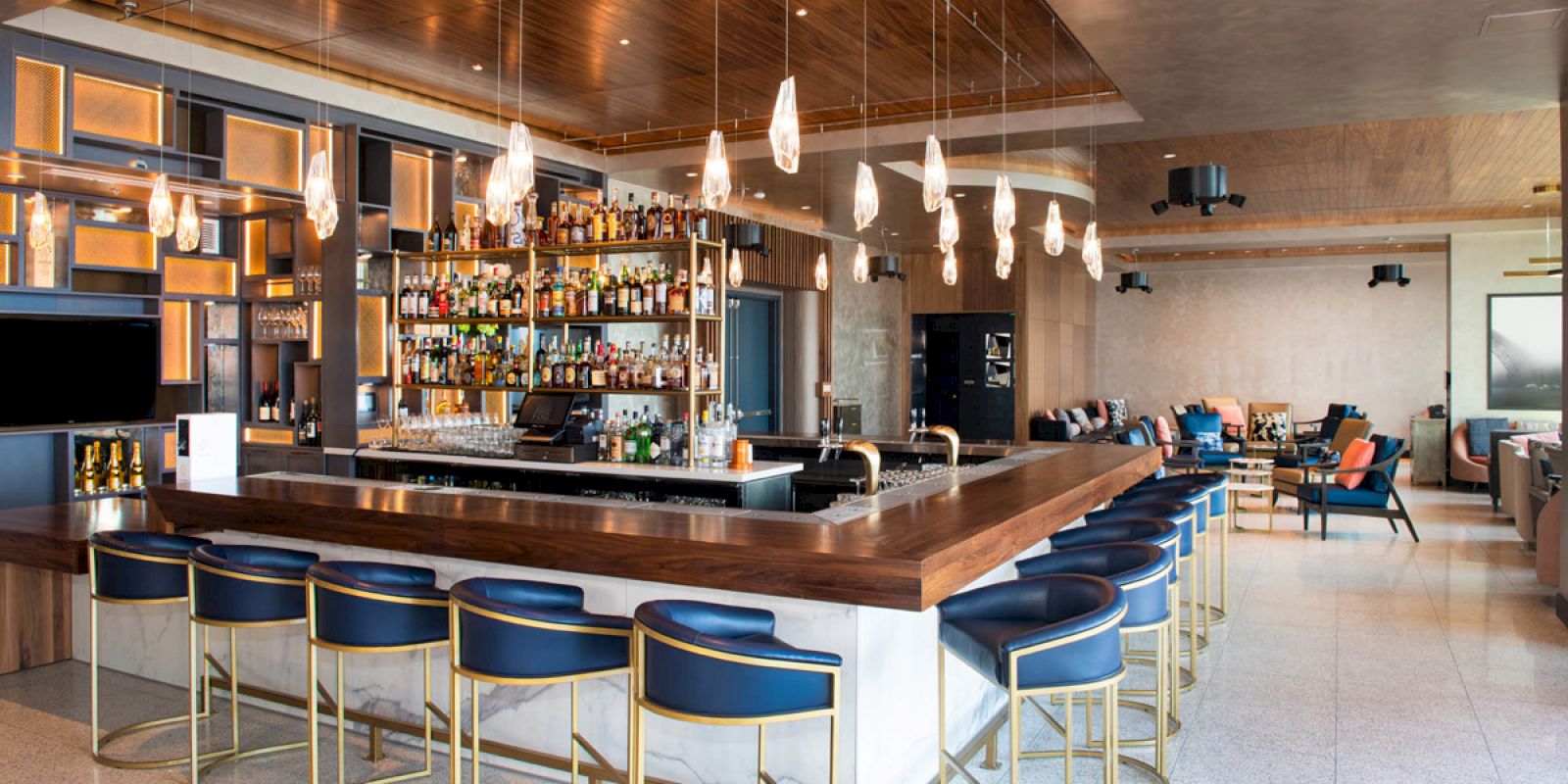 The image shows a modern bar with a wooden counter, high-back chairs, shelves stocked with liquor, pendant lights, and seating areas in the background.