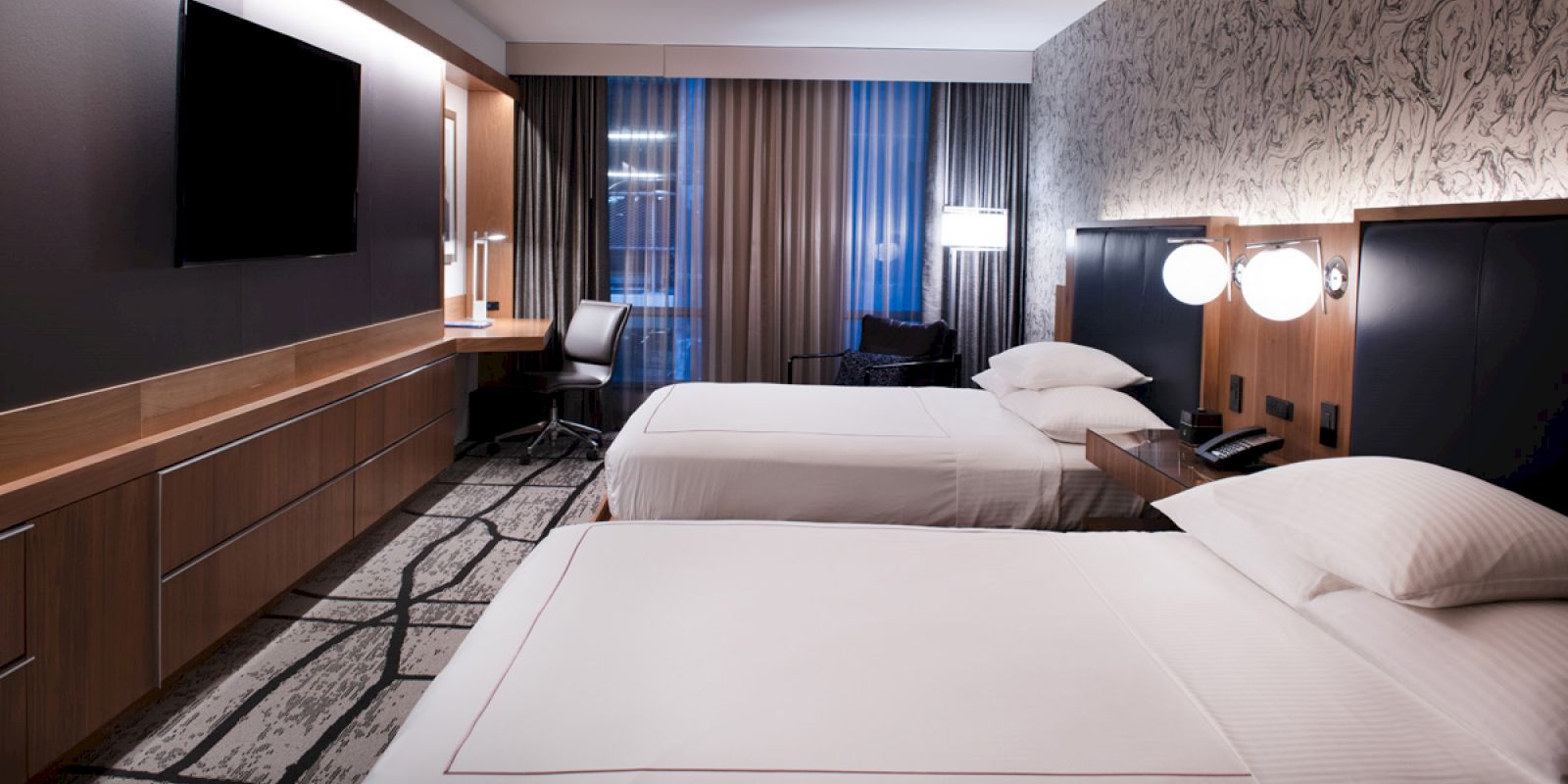This image shows a modern hotel room with two neatly made beds, a flat-screen TV, a work desk and chair, and contemporary decor with patterned wallpaper.