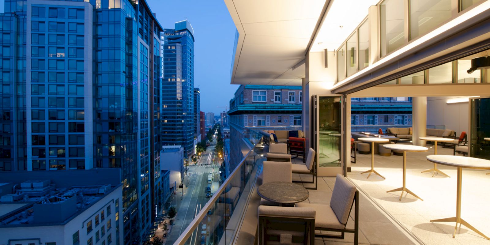 A modern balcony overlooks a cityscape with tall buildings. The balcony features tables, chairs, and is connected to an indoor area. It's dusk.