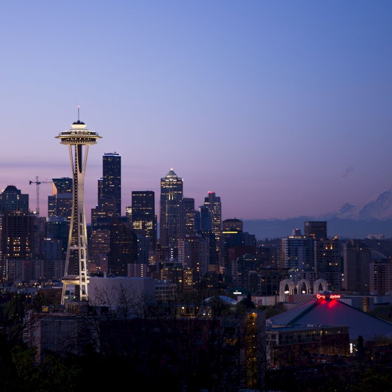 Skyline of a city with the Space Needle prominently featured, against a twilight sky with a mountain in the background.