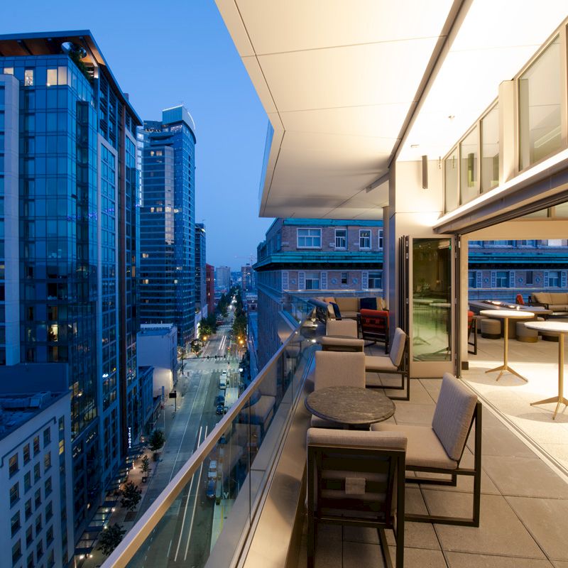 An urban balcony view at dusk, featuring modern buildings and a well-lit outdoor seating area with tables and chairs.