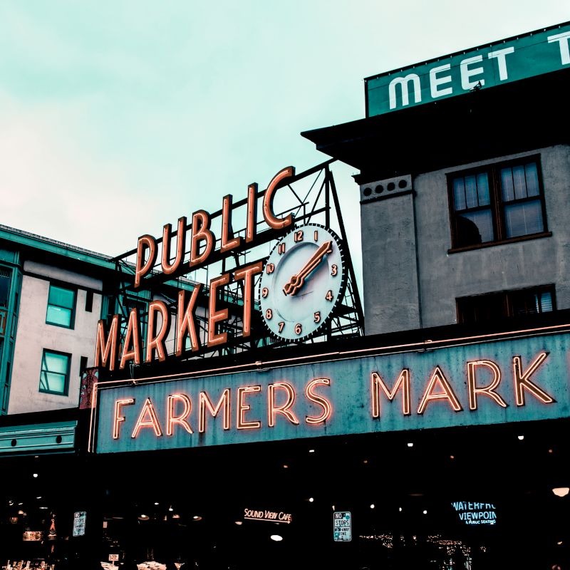 The image shows a large public market with a clock, neon signs reading "Public Market" and "Farmers Market," and nearby buildings in the background.