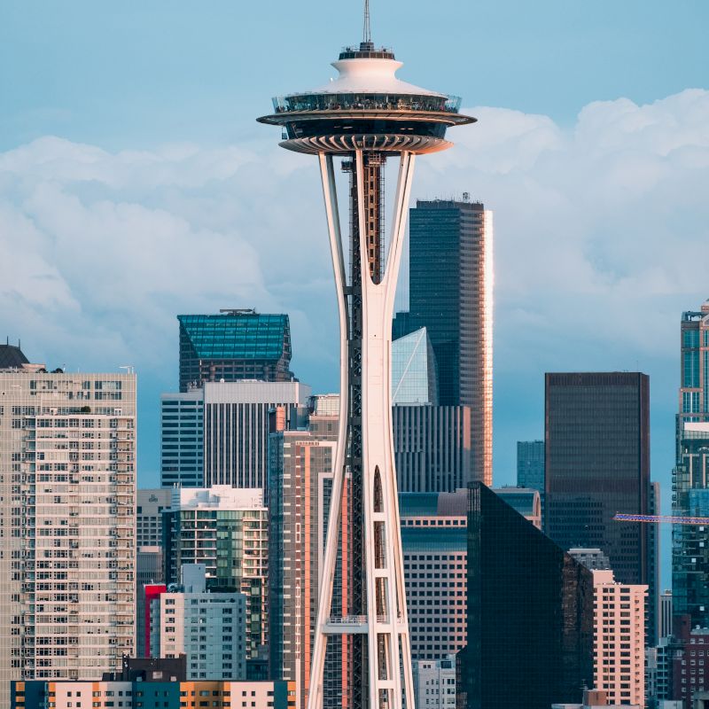 The image shows the Space Needle in Seattle, surrounded by modern skyscrapers and buildings, against a backdrop of clouds and blue sky.